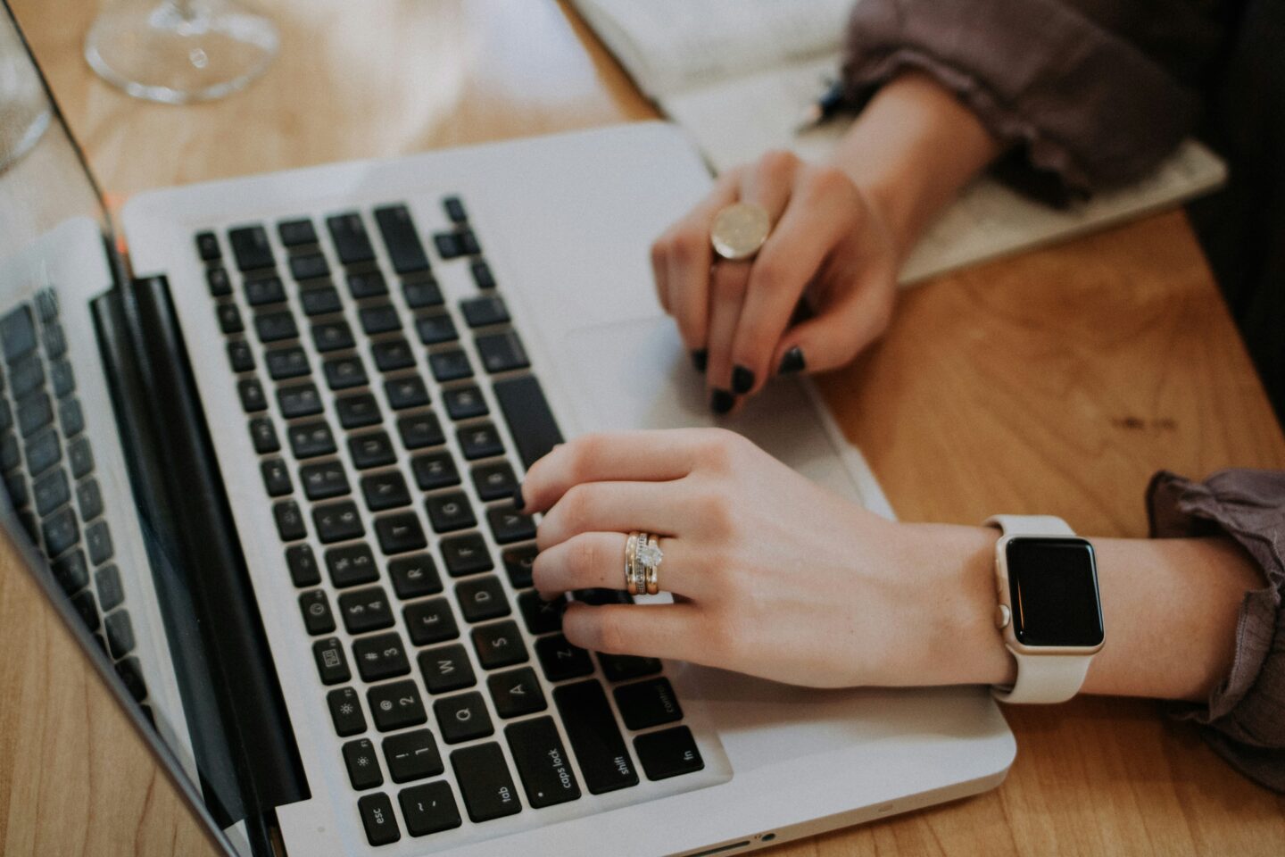 Close-up of hands typing on a laptop keyboard, wearing rings and a smartwatch, with a wooden desk and glass nearby.