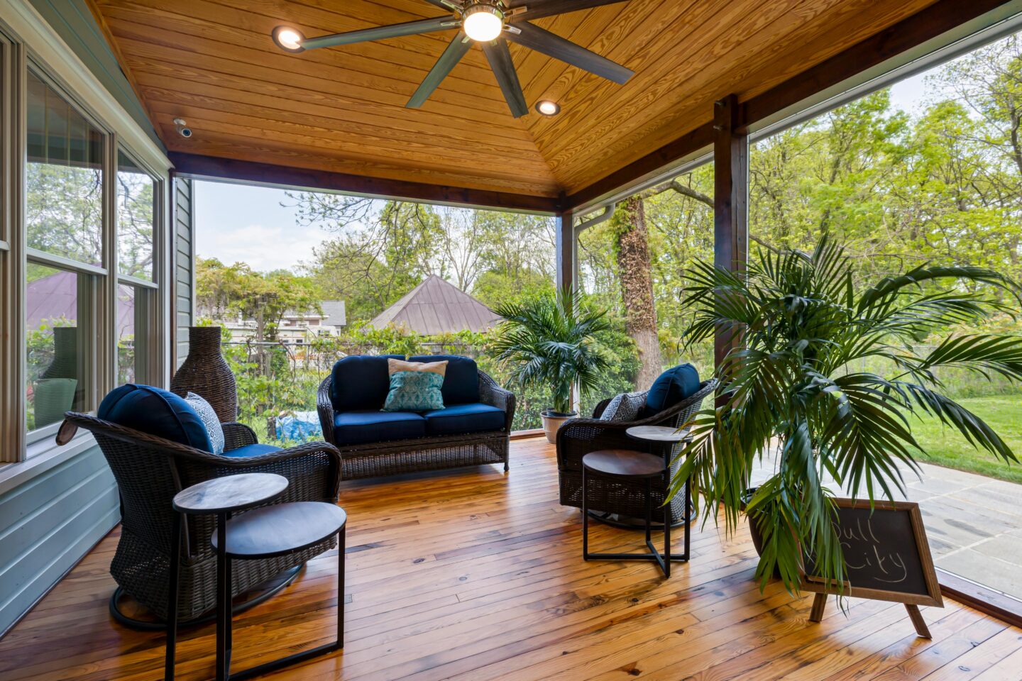 Modern screened-in patio with vaulted wood ceiling, ceiling fan, wicker seating with navy cushions, indoor plants, and polished hardwood flooring overlooking a lush backyard garden.