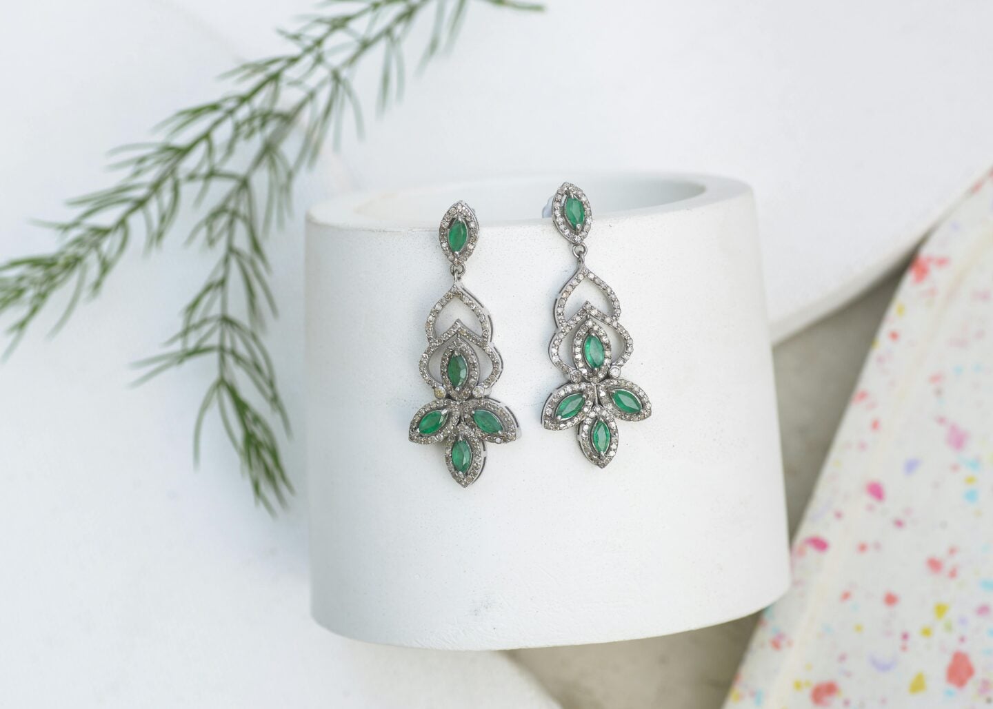 Ornate silver drop earrings with green gemstones and crystal detailing displayed on a white stand, elegant jewelry for weddings or formal occasions.