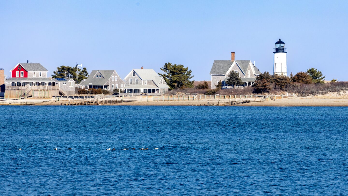 Scenic Cape Cod lighthouse by the sea with coastal homes and calm blue water along the Massachusetts shoreline.