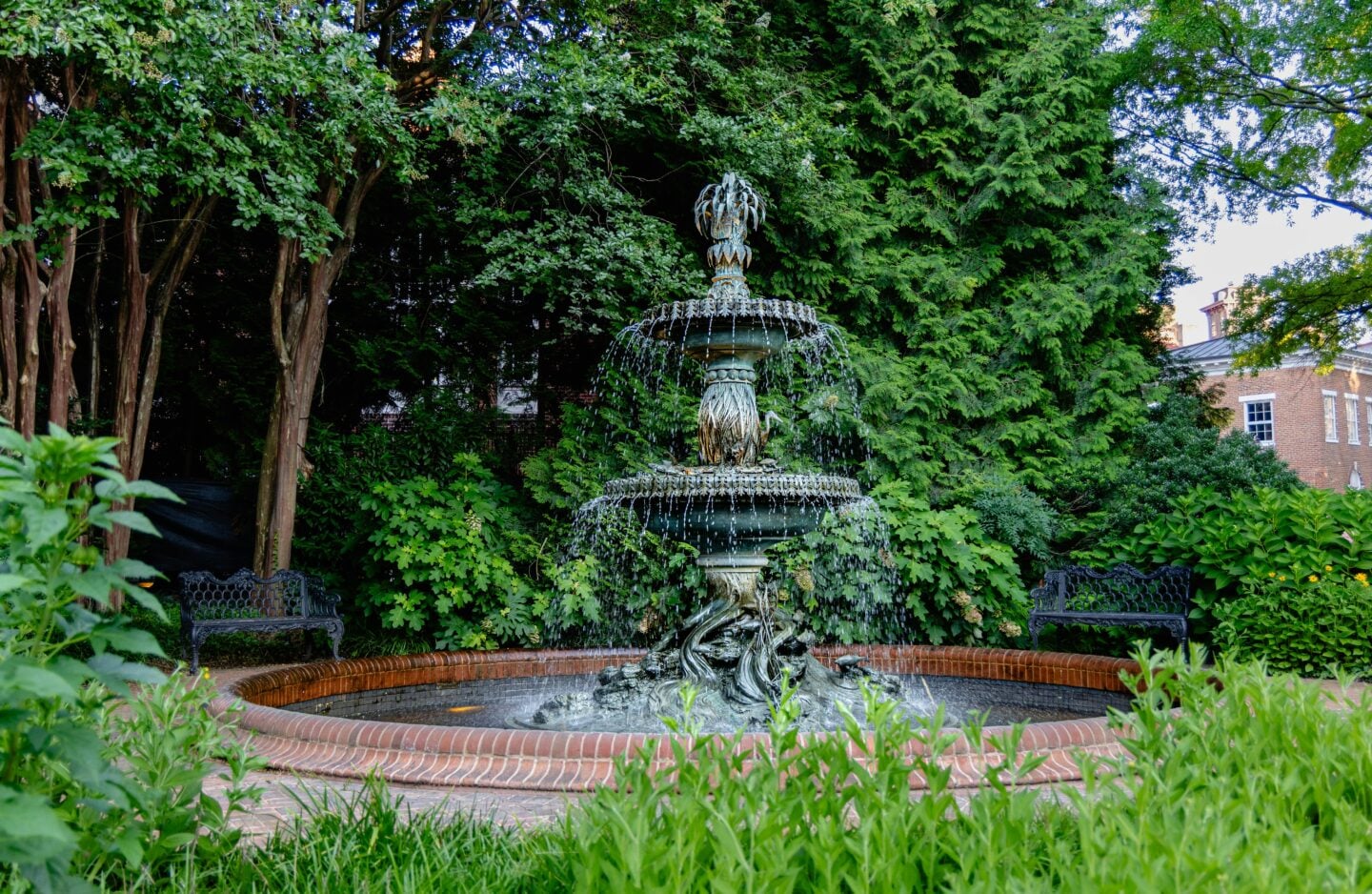 Ornate stone garden fountain surrounded by lush greenery, brick edging, and shaded trees in a peaceful outdoor setting.