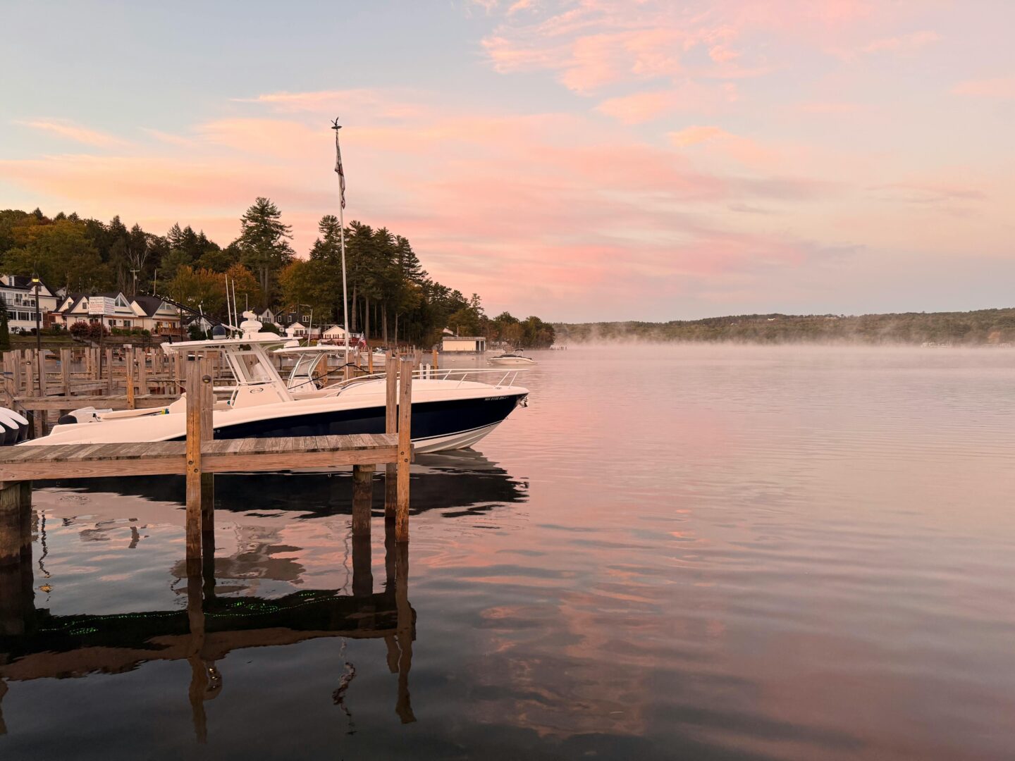 Serene sunset at Lake Winnipesaukee dock in New Hampshire with boat moored at wooden pier and soft pink sky reflecting over calm water.