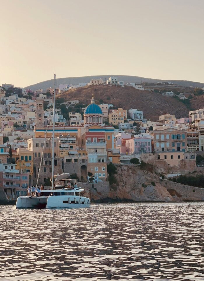Colorful neoclassical buildings and a blue-domed church overlooking the sea in Syros, Greece, with a catamaran anchored in the harbor at sunset.