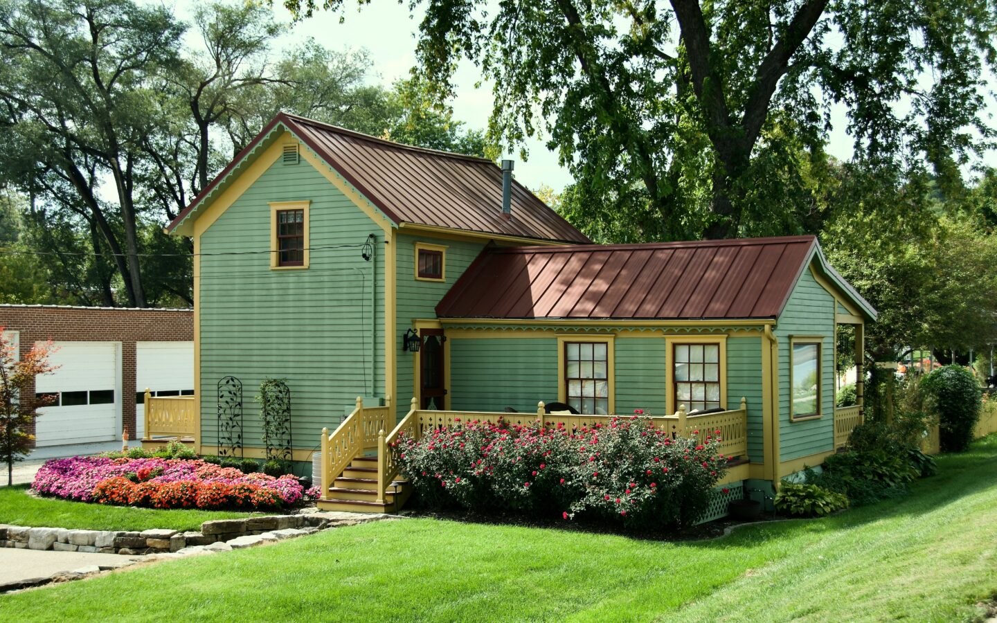 Small green house with yellow trim surrounded by a landscaped garden and colorful flowers on a sunny day.