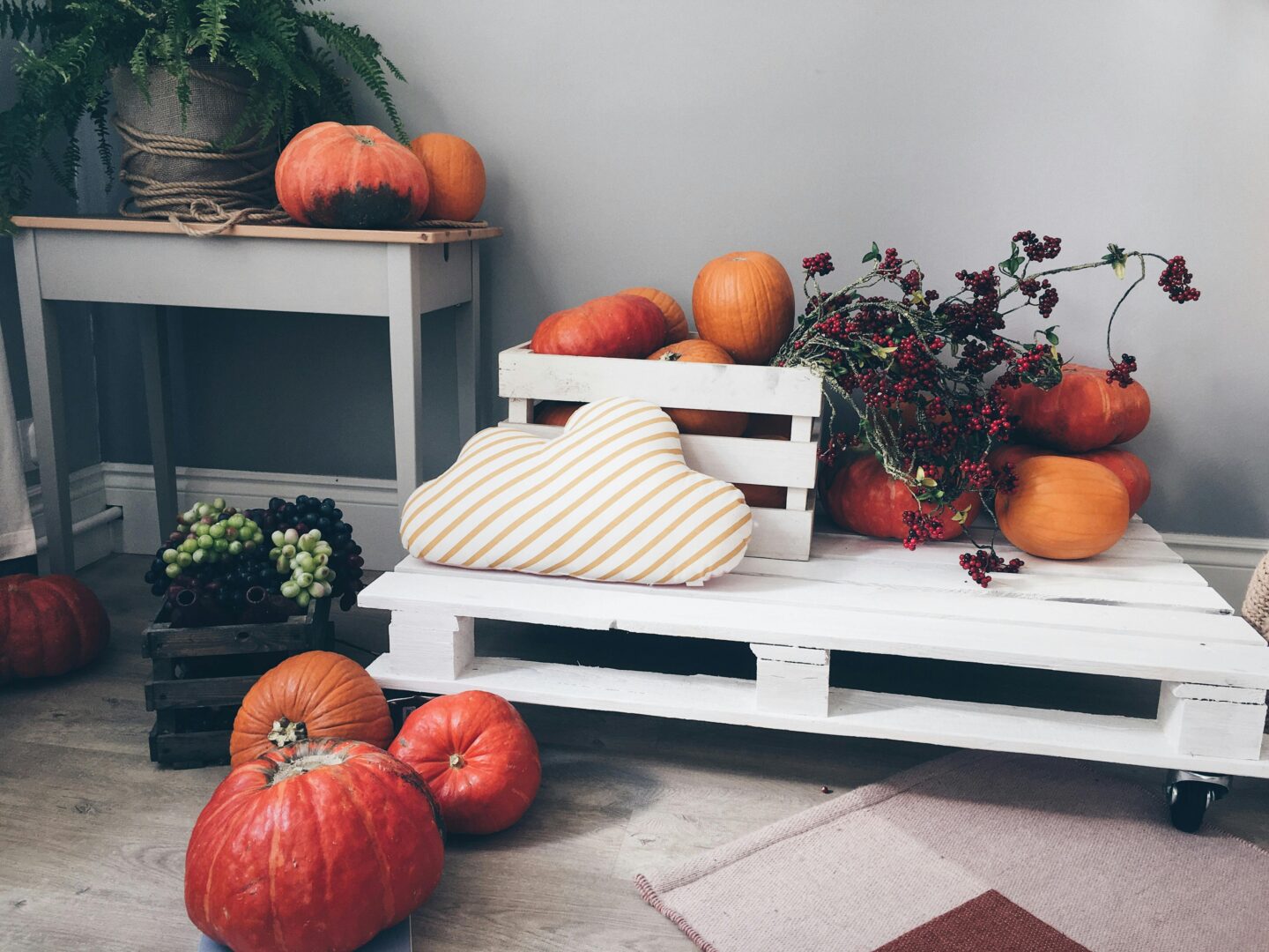 Autumn home decor with pumpkins, red berries, and grapes arranged on a white wooden pallet table with a striped cloud-shaped pillow indoors.