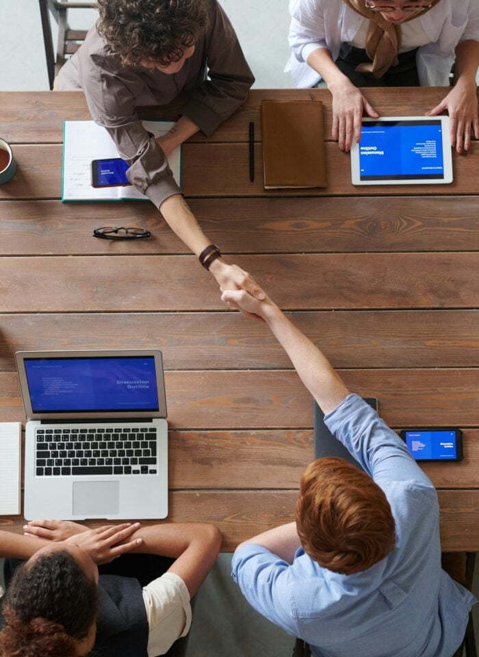 Team members shaking hands across a wooden meeting table during a collaborative business meeting with laptops and tablets.