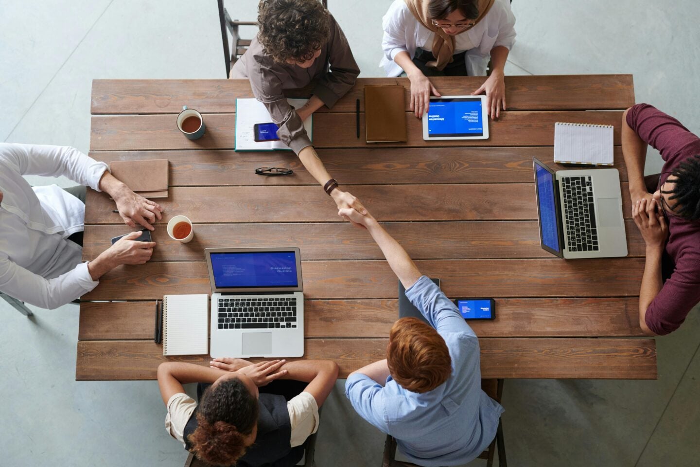 Team members shaking hands across a wooden meeting table during a collaborative business meeting with laptops and tablets.