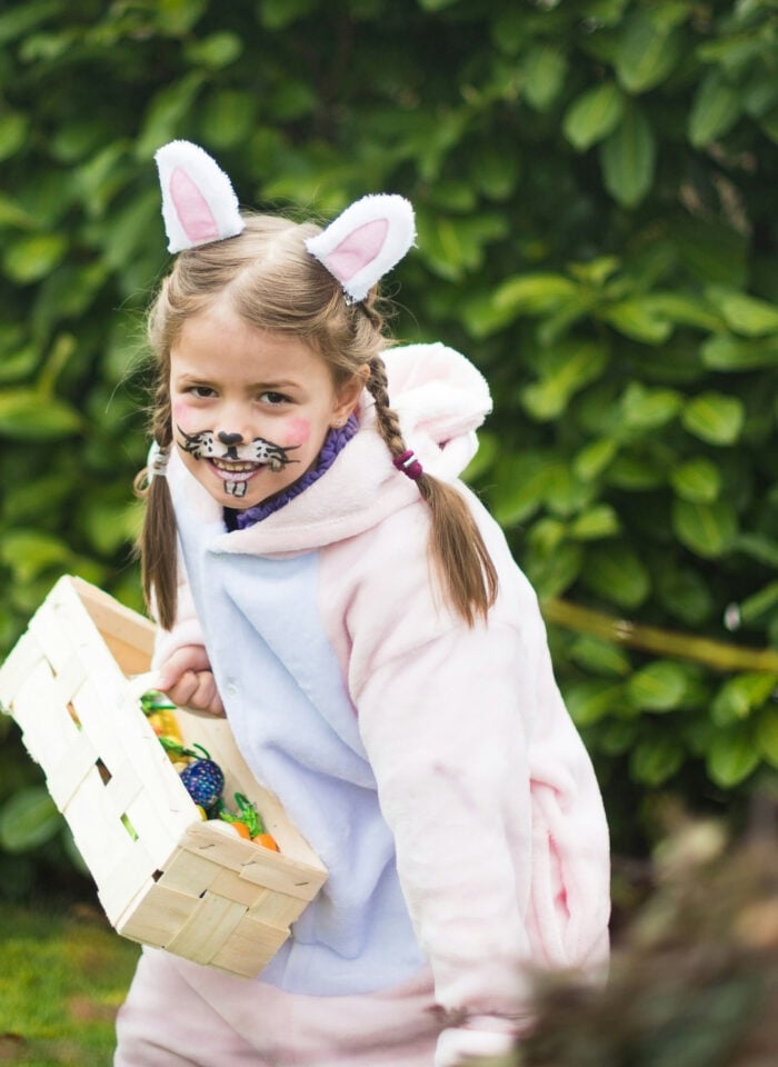 Young girl dressed in a bunny costume with face paint, holding a basket during an Easter egg hunt outdoors in a garden surrounded by greenery.