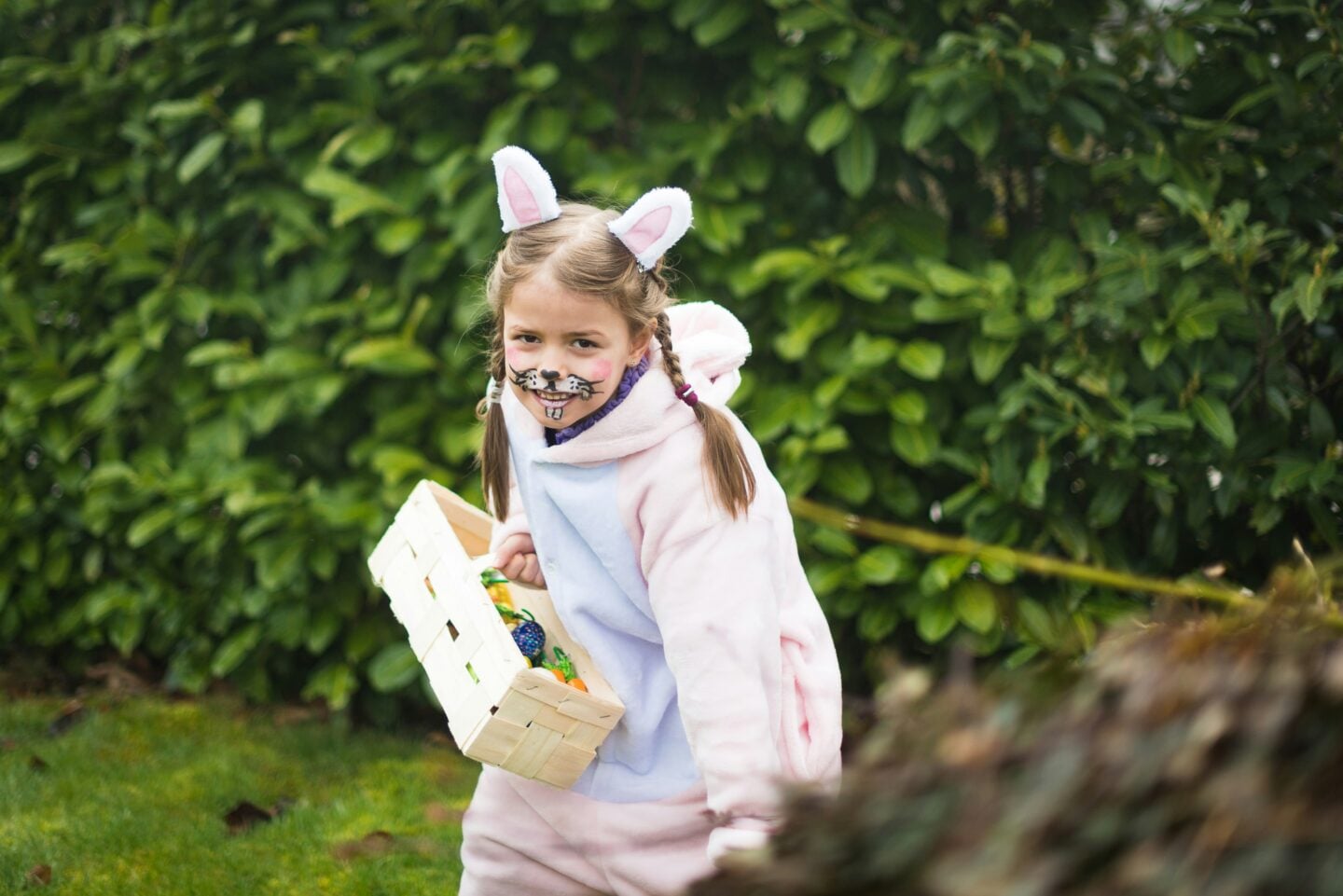 Young girl dressed in a bunny costume with face paint, holding a basket during an Easter egg hunt outdoors in a garden surrounded by greenery.