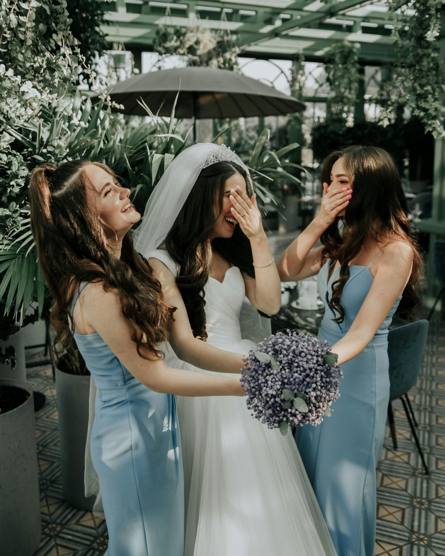 Bride in white wedding dress laughing with bridesmaids in light blue dresses, holding bouquet in lush greenhouse setting.