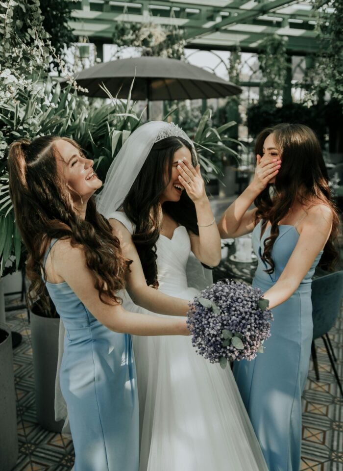 Bride in white wedding dress laughing with bridesmaids in light blue dresses, holding bouquet in lush greenhouse setting.