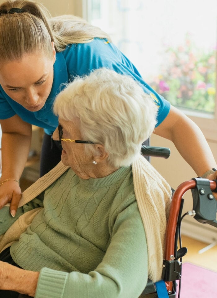 Caregiver in blue uniform assisting an elderly woman sitting in a wheelchair at home, leaning in to speak while adjusting a blanket over her shoulders.