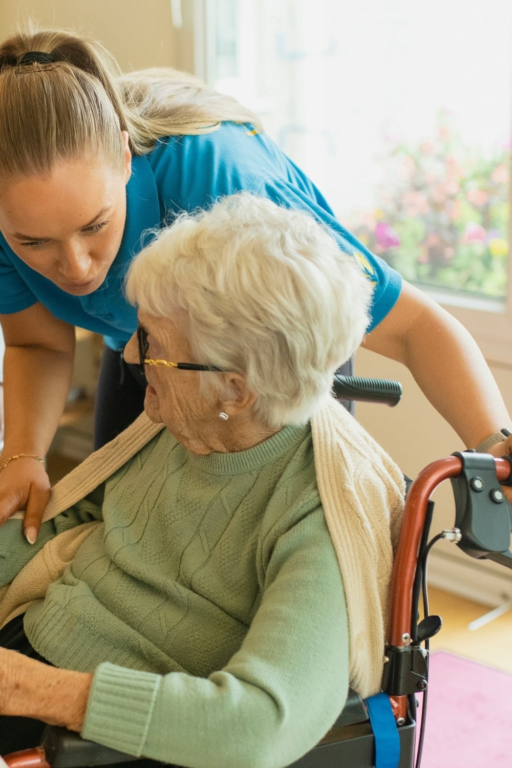 Caregiver in blue uniform assisting an elderly woman sitting in a wheelchair at home, leaning in to speak while adjusting a blanket over her shoulders.