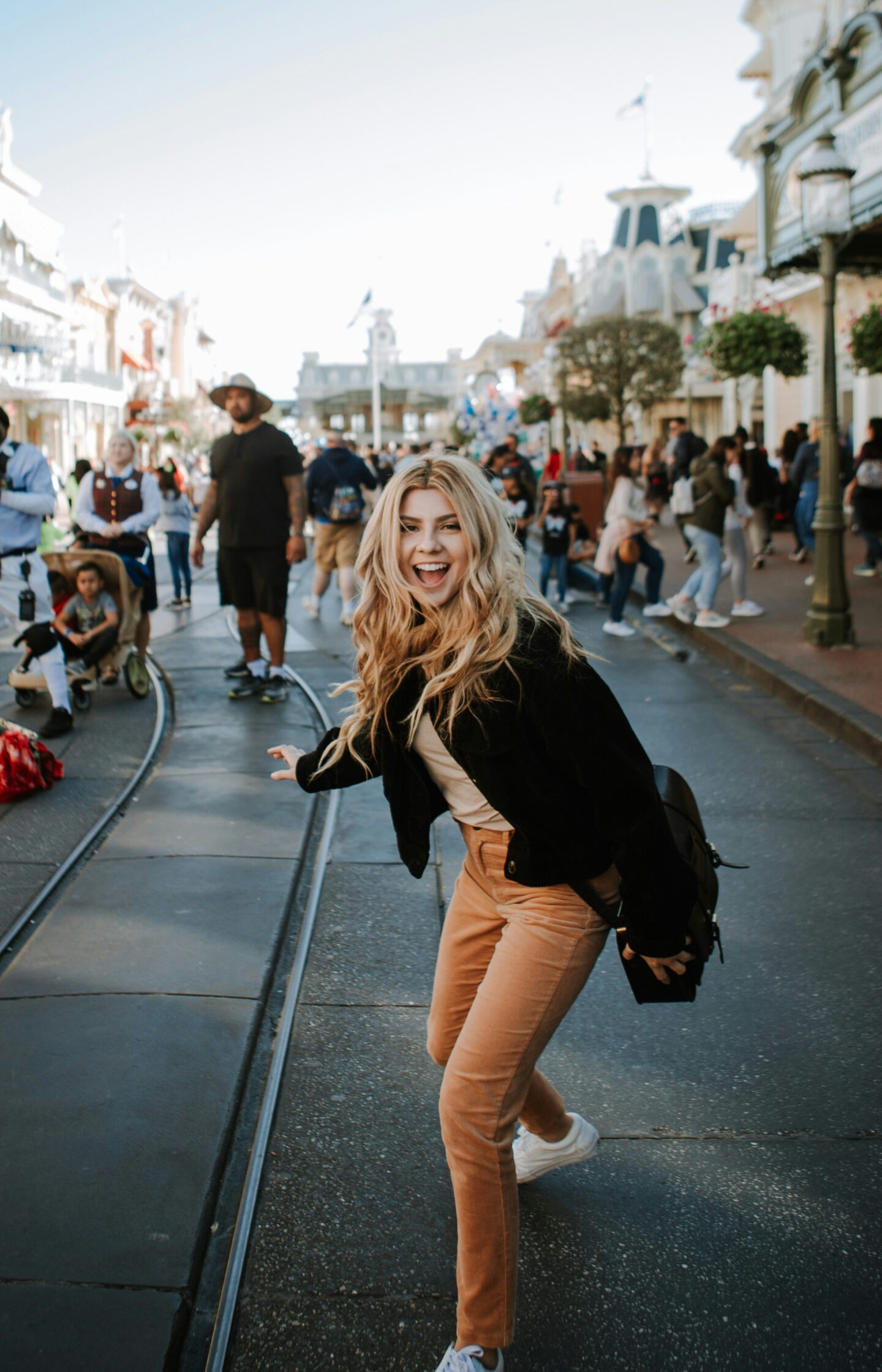 Smiling woman with voluminous blonde hair posing playfully on a busy theme park street, confident female traveler in casual outfit with beige pants and black jacket, lively crowd and sunny outdoor atmosphere.