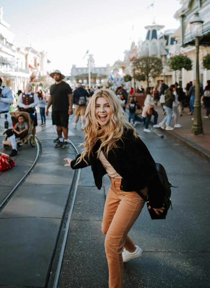 Smiling woman with voluminous blonde hair posing playfully on a busy theme park street, confident female traveler in casual outfit with beige pants and black jacket, lively crowd and sunny outdoor atmosphere.