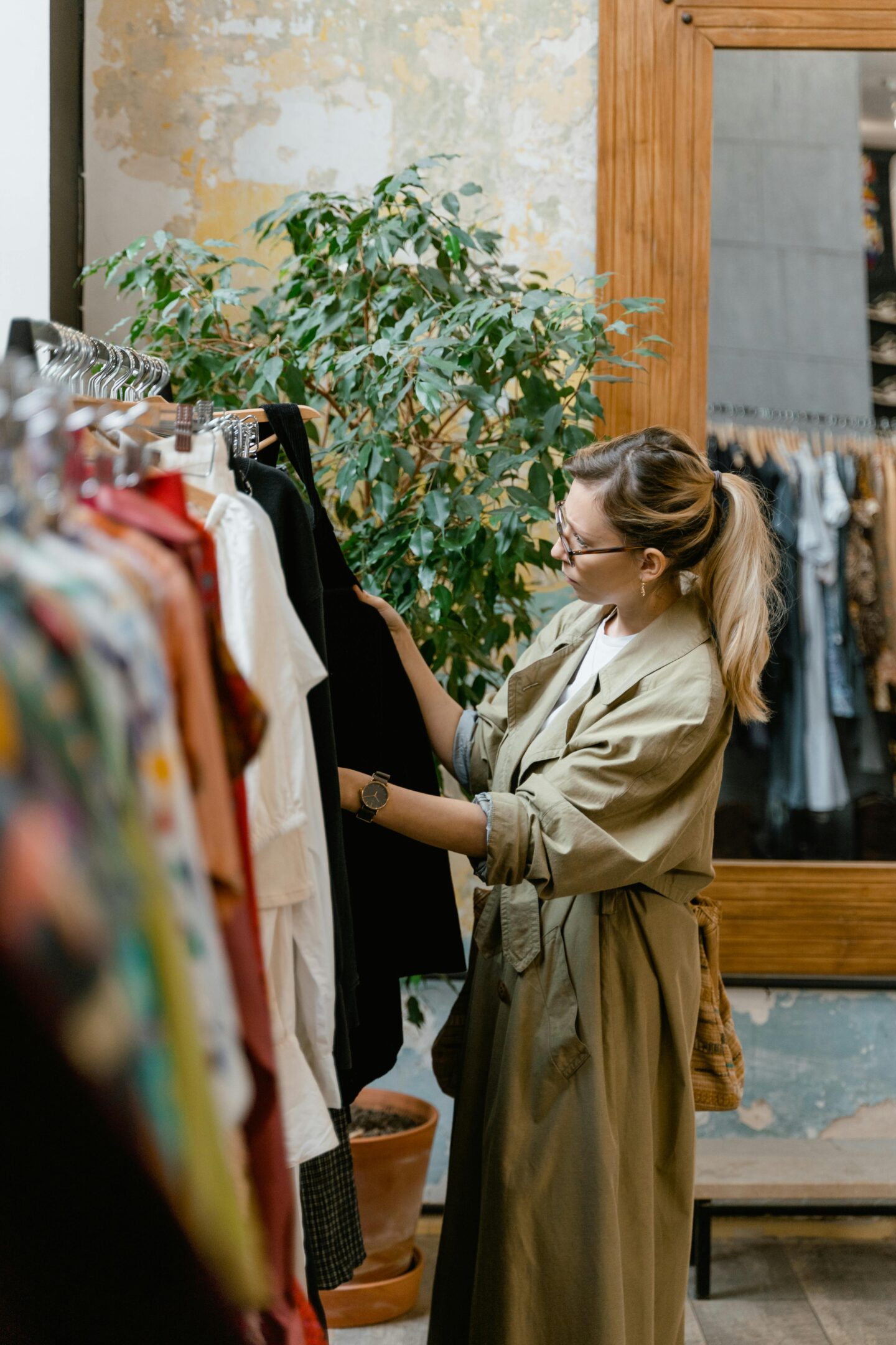 Woman browsing clothing rack in boutique store, selecting outfit, stylish shopper in trench coat, indoor retail fashion scene with plants and mirror, warm natural tones.