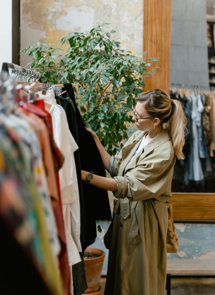 Woman browsing clothing rack in boutique store, selecting outfit, stylish shopper in trench coat, indoor retail fashion scene with plants and mirror, warm natural tones.
