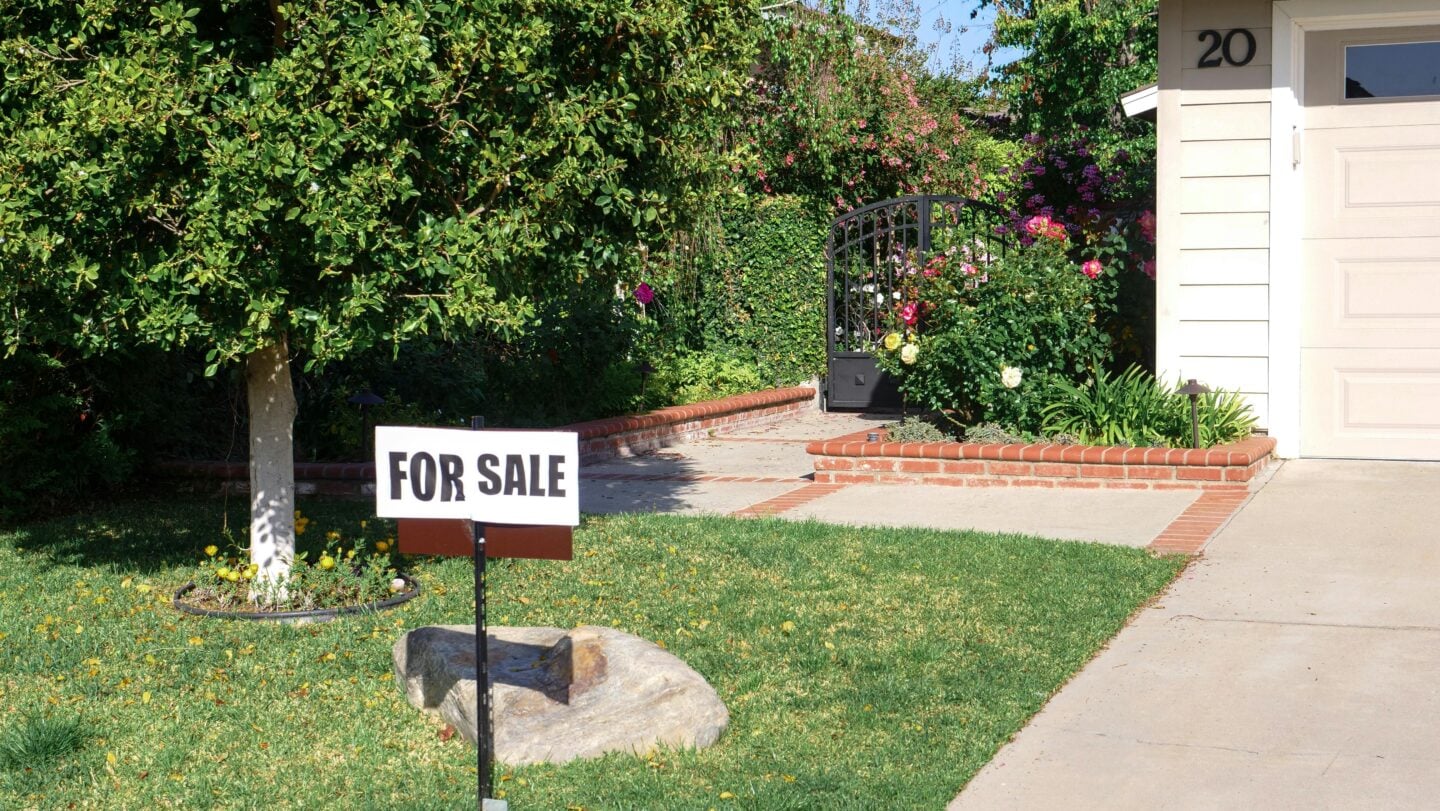 House with a for sale sign in the front yard and landscaped garden in a residential neighborhood.