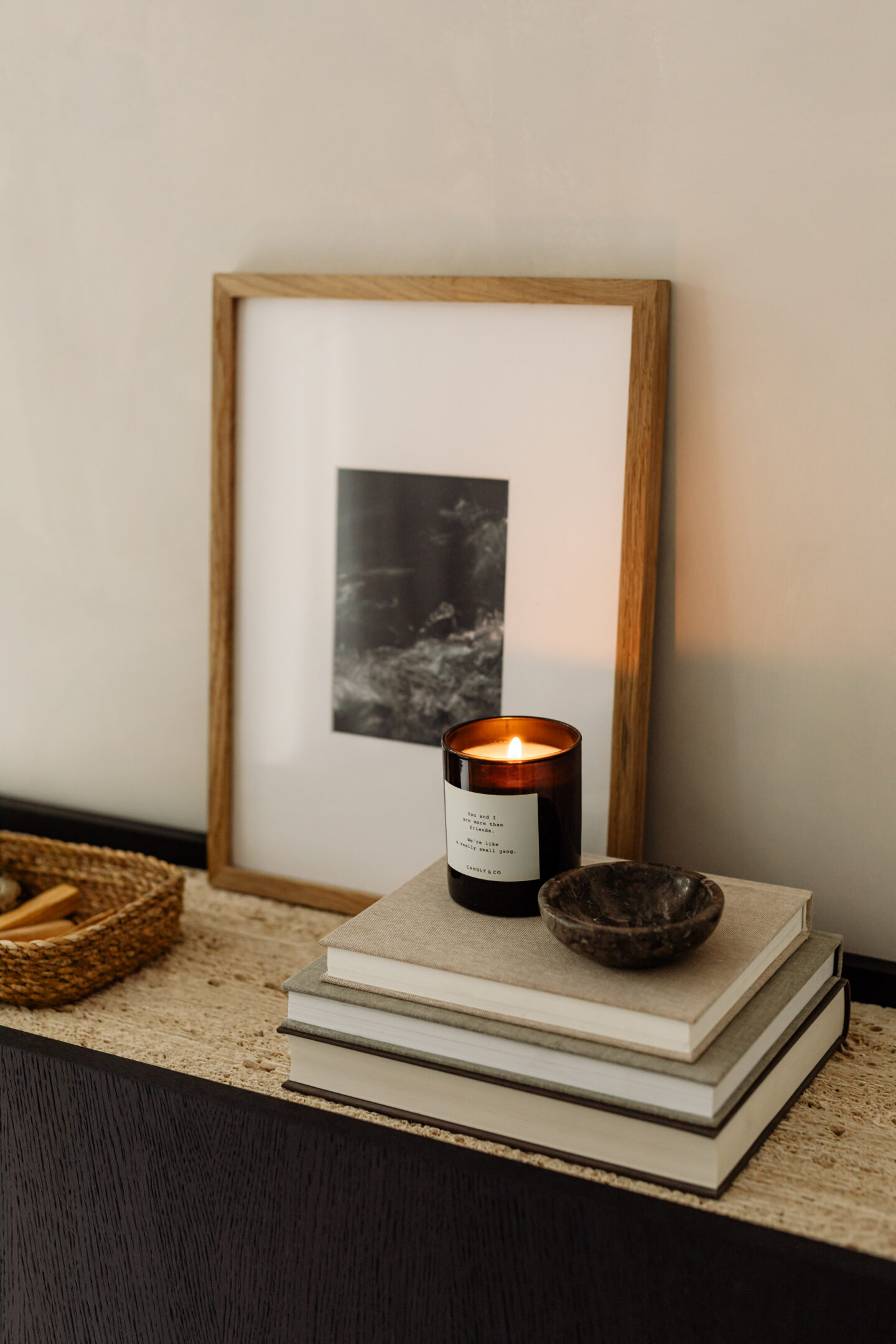 Styled home decor with lit candle on stacked books, framed artwork, and woven tray on wooden console table