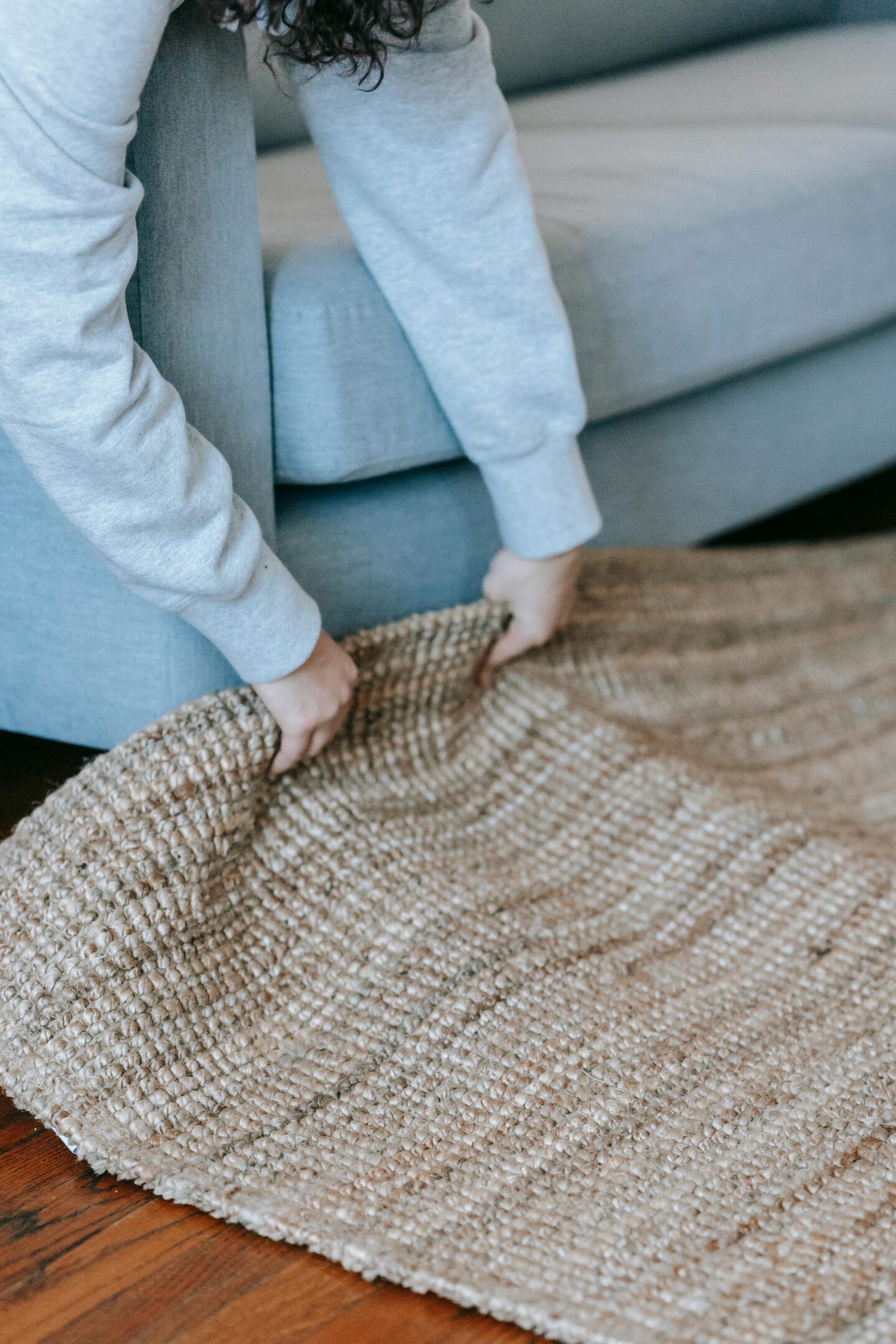 Person adjusting textured jute rug on wooden floor next to light gray sofa in cozy living room