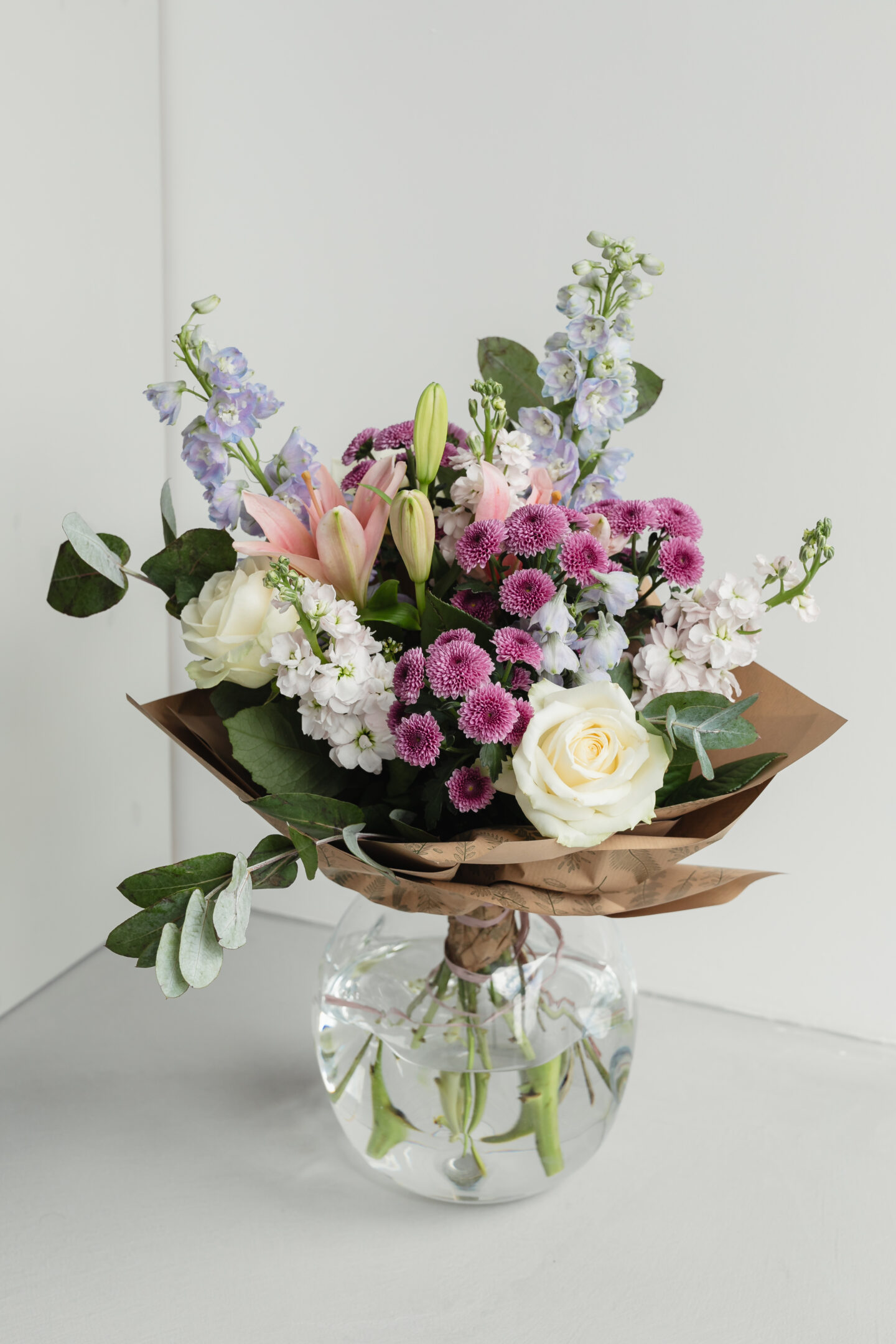 Elegant mixed flower bouquet with white roses, pink lilies, purple chrysanthemums, and light blue delphiniums arranged in brown paper and placed in a clear glass vase on a table.