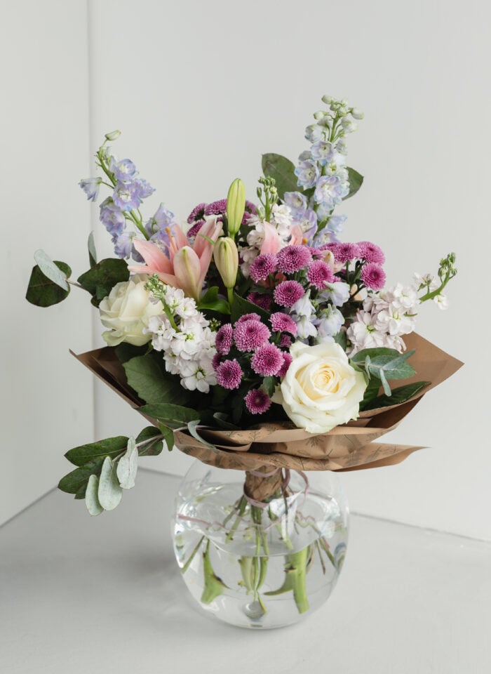 Elegant mixed flower bouquet with white roses, pink lilies, purple chrysanthemums, and light blue delphiniums arranged in brown paper and placed in a clear glass vase on a table.