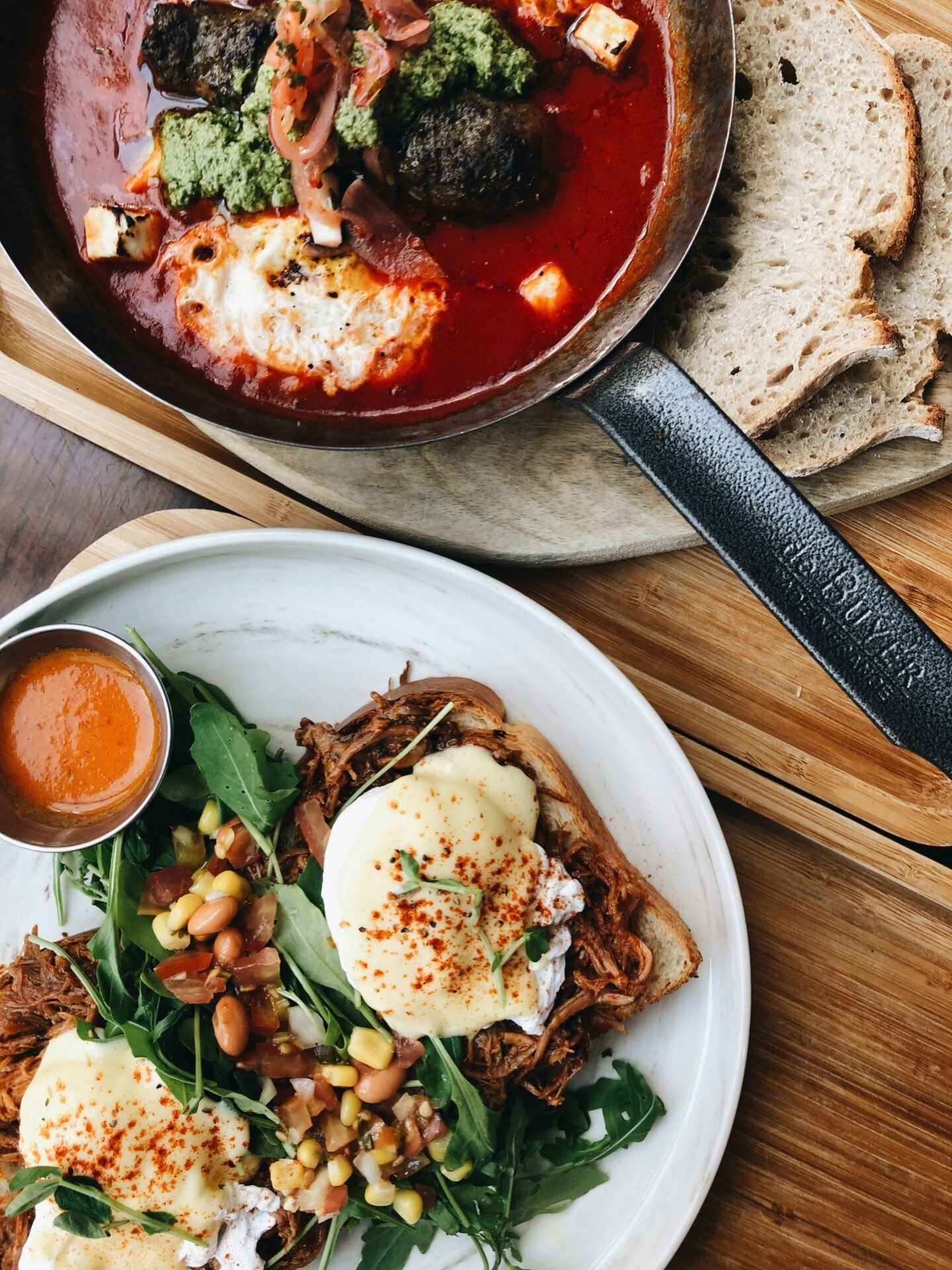 Gourmet brunch plate with poached eggs, pulled meat on toast, fresh greens, corn salad, and sauce served on a white plate.