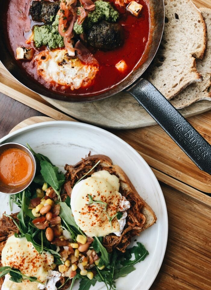 Gourmet brunch plate with poached eggs, pulled meat on toast, fresh greens, corn salad, and sauce served on a white plate.