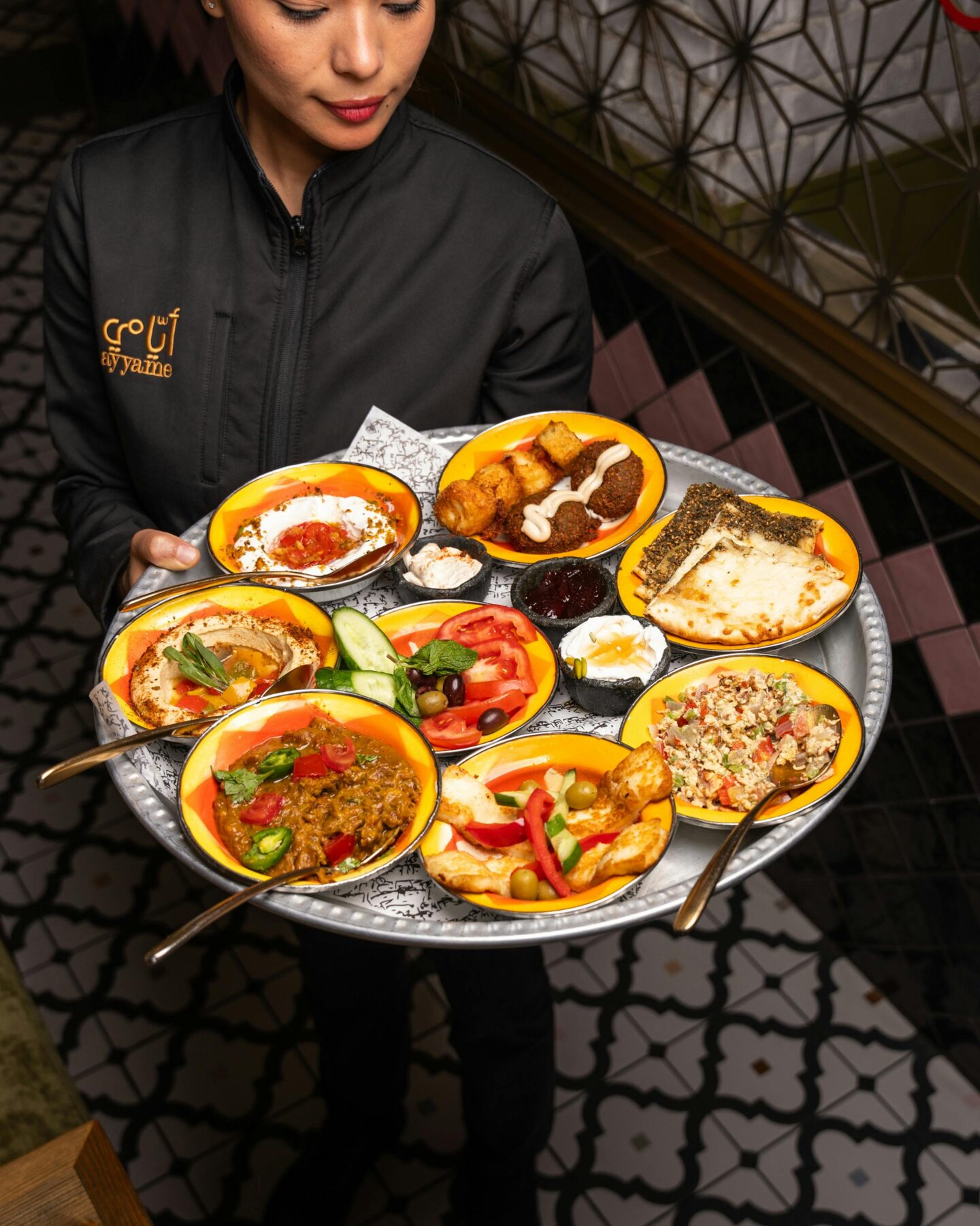 Server carrying a tray of colorful Middle Eastern mezze dishes including hummus, falafel, vegetables, dips, and flatbread.