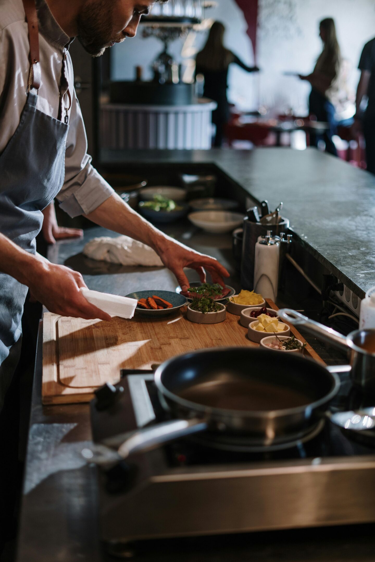 Chef preparing ingredients on a wooden cutting board in a restaurant kitchen with pans and cooking tools nearby.