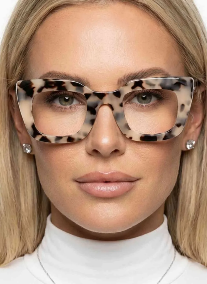 Close-up portrait of woman wearing oversized tortoiseshell eyeglasses, blonde hair and neutral makeup, stylish accessories with minimal white background.