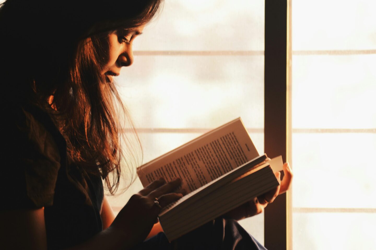 Woman reading a book by a window in warm sunlight during a relaxing travel moment.