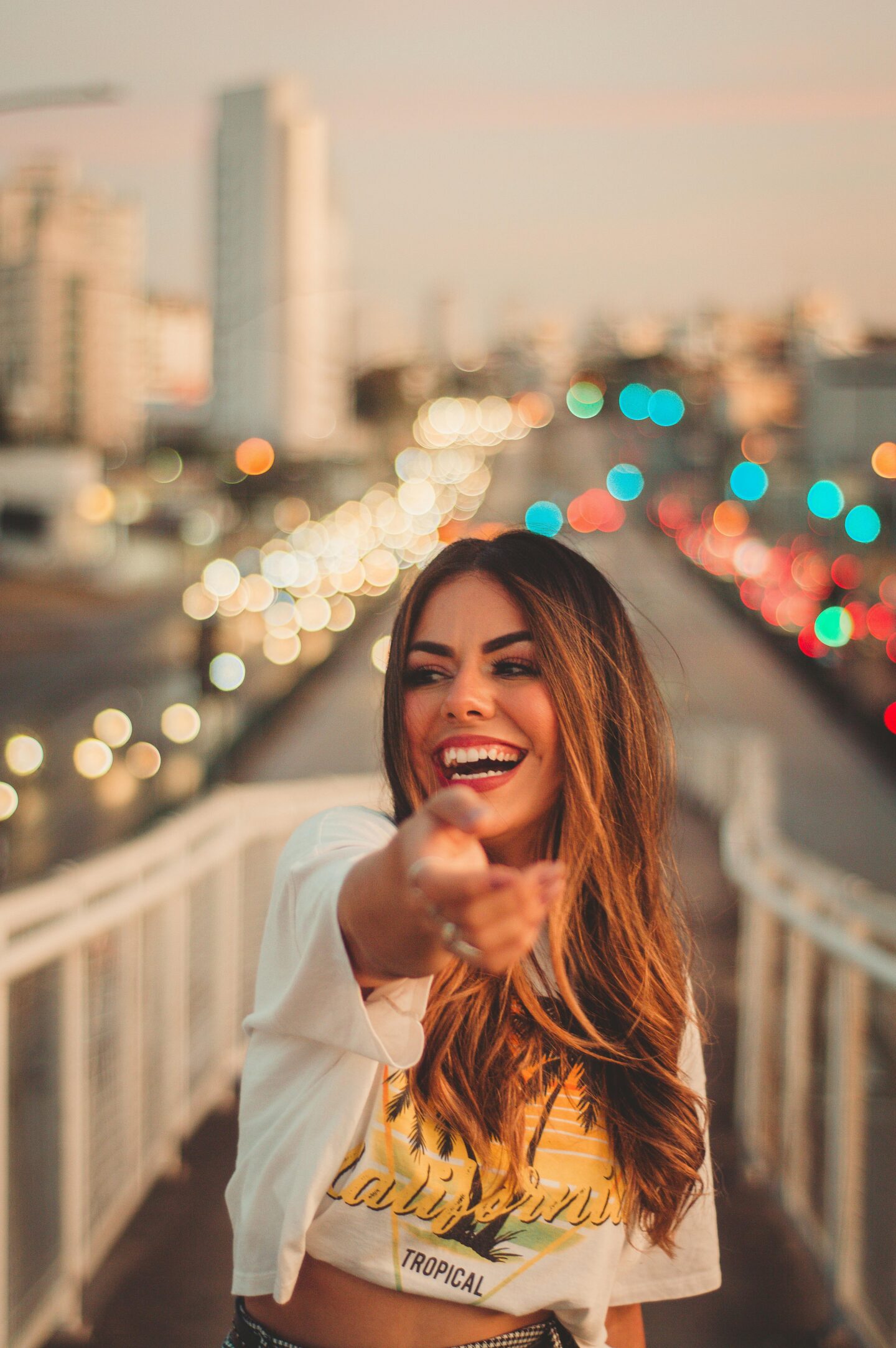 Smiling woman pointing toward the camera while standing on a pedestrian bridge with blurred city traffic lights in the background.