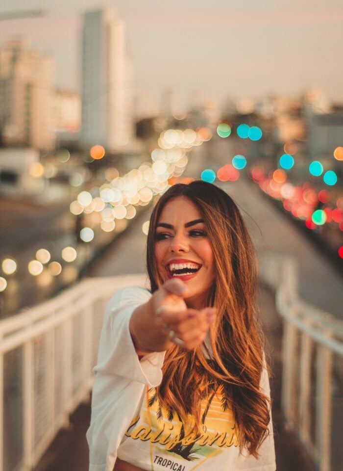 Smiling woman pointing toward the camera while standing on a pedestrian bridge with blurred city traffic lights in the background.