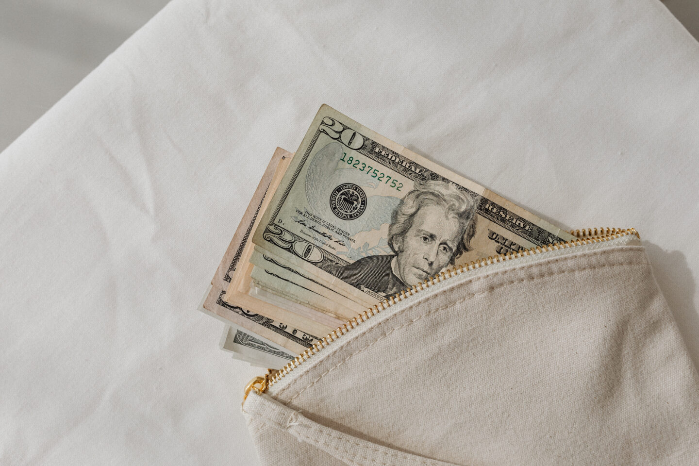 US dollar bills partially visible inside a small zip pouch on a white fabric surface.