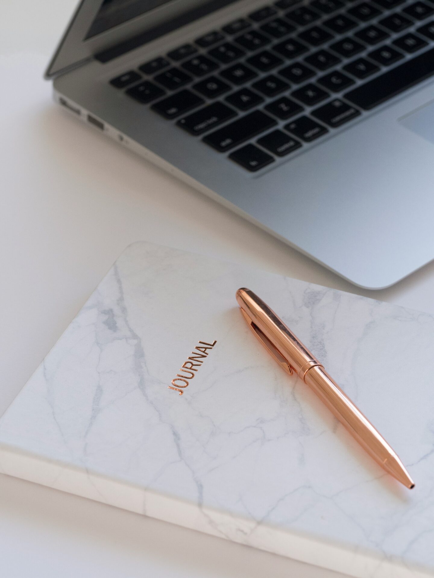 Minimal desk setup with a marble notebook labeled journal, a rose gold pen, and a laptop.