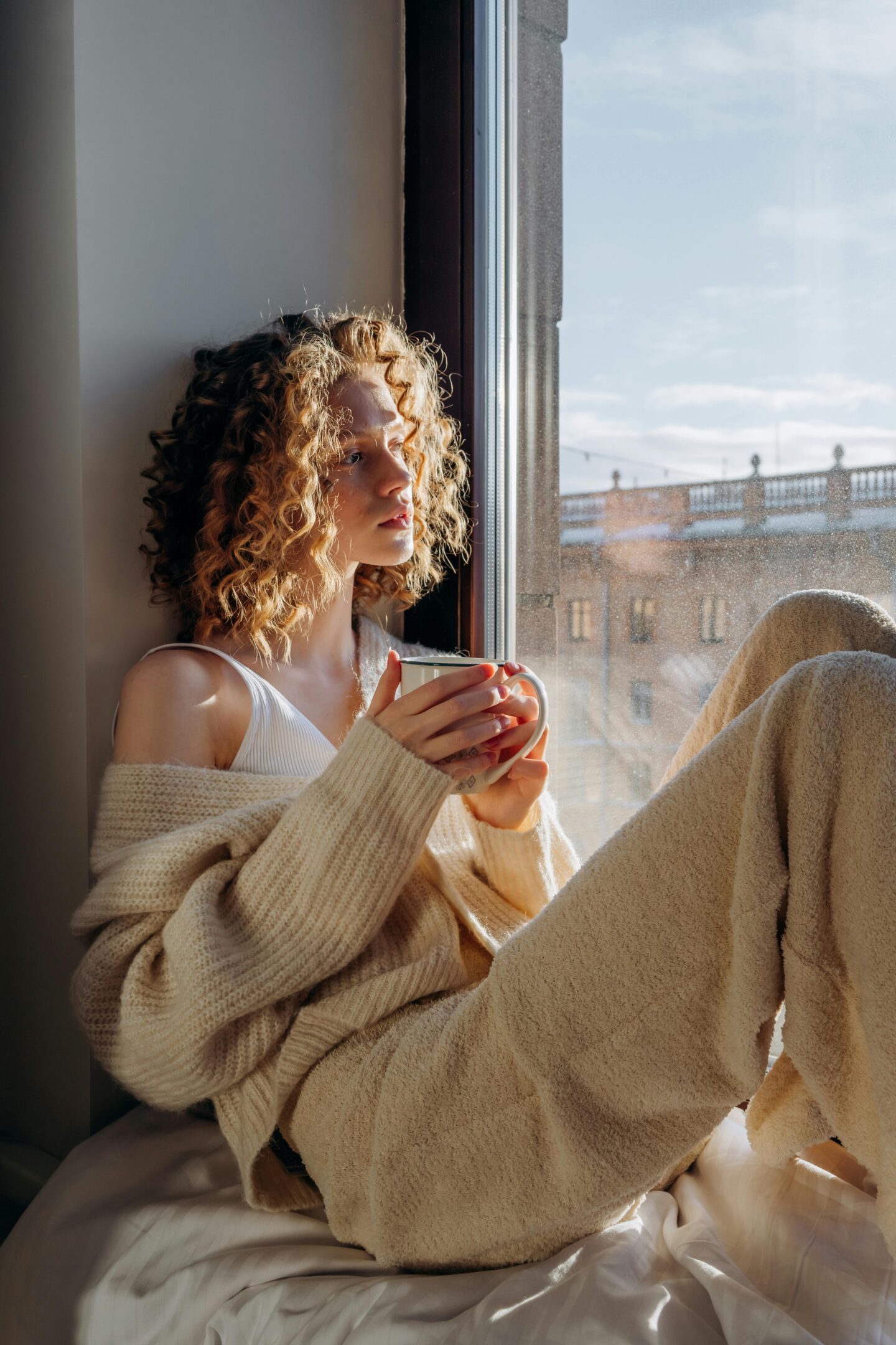 Young woman sitting by a window in cozy clothes holding a mug and enjoying morning sunlight.
