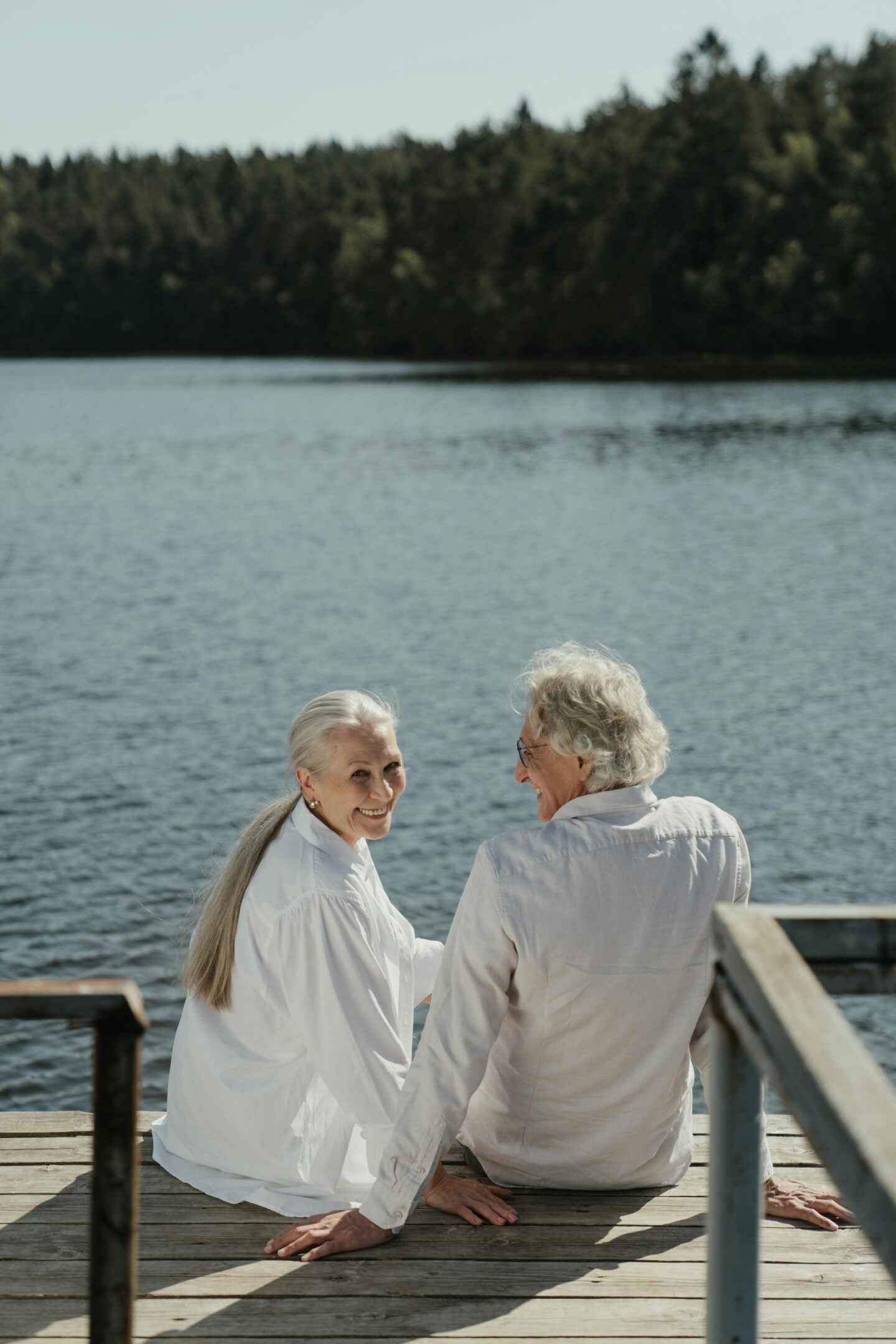Senior couple sitting on a wooden dock by a lake, smiling and holding hands while enjoying a peaceful retirement moment surrounded by forested scenery.