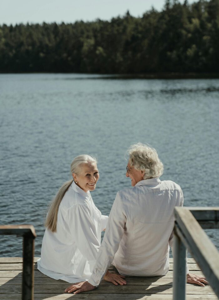 Senior couple sitting on a wooden dock by a lake, smiling and holding hands while enjoying a peaceful retirement moment surrounded by forested scenery