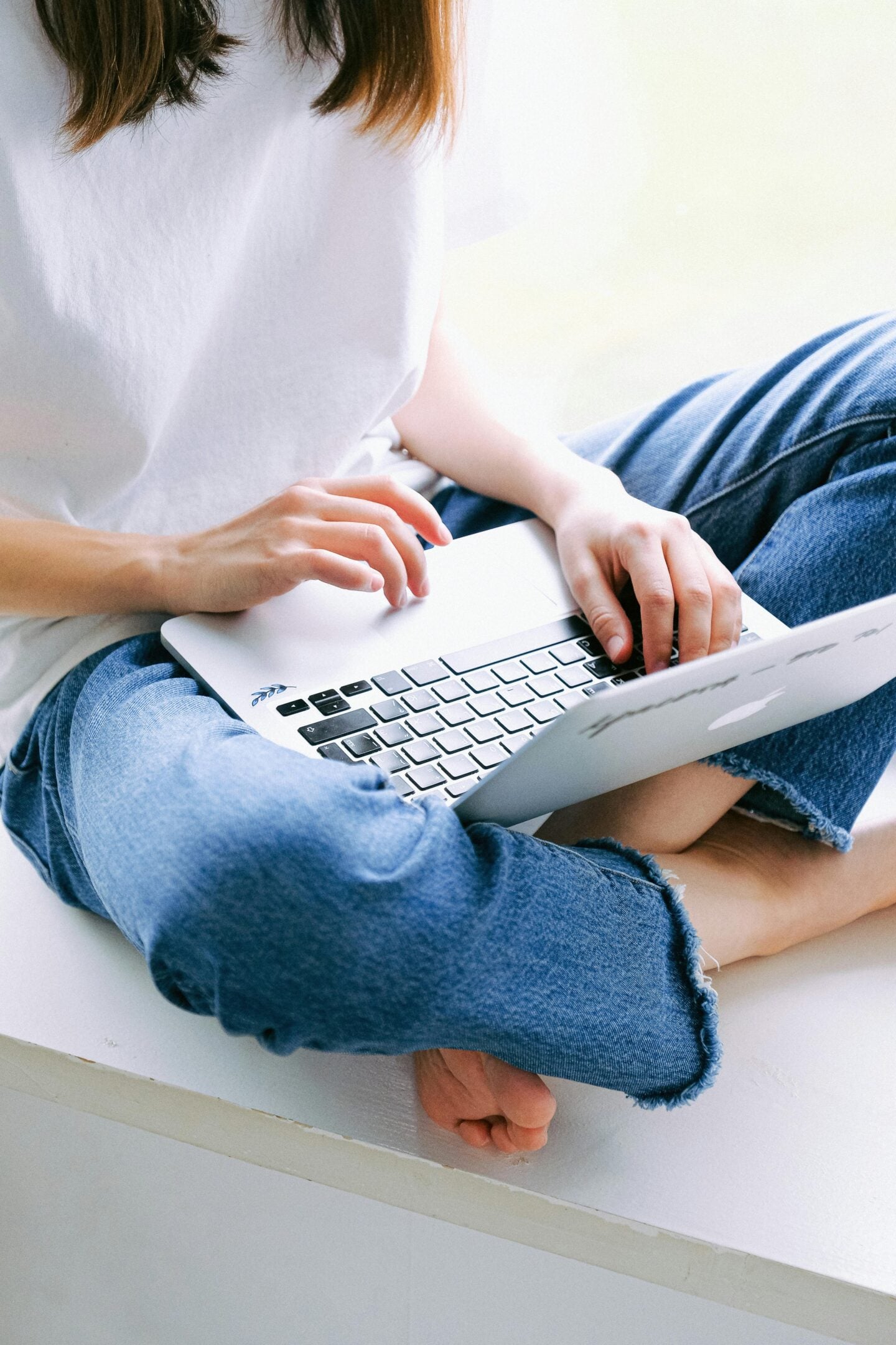 Person sitting cross-legged in jeans using a laptop, typing on the keyboard in a bright, minimal indoor setting.