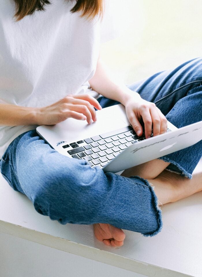 Person sitting cross-legged in jeans using a laptop, typing on the keyboard in a bright, minimal indoor setting.