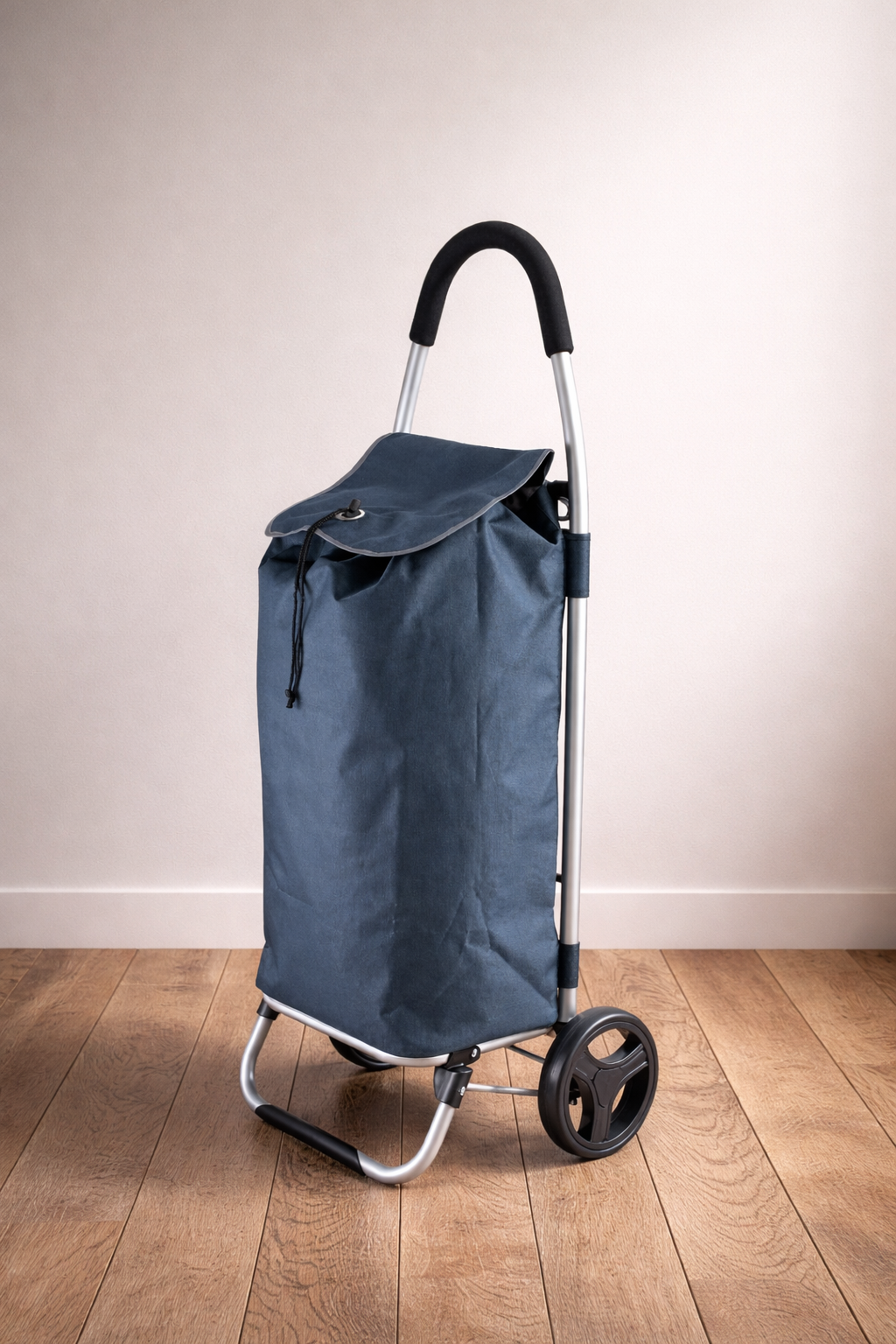 A modern folding shopping trolley with a dark blue fabric bag and silver metal frame standing on a wooden floor against a plain beige wall.