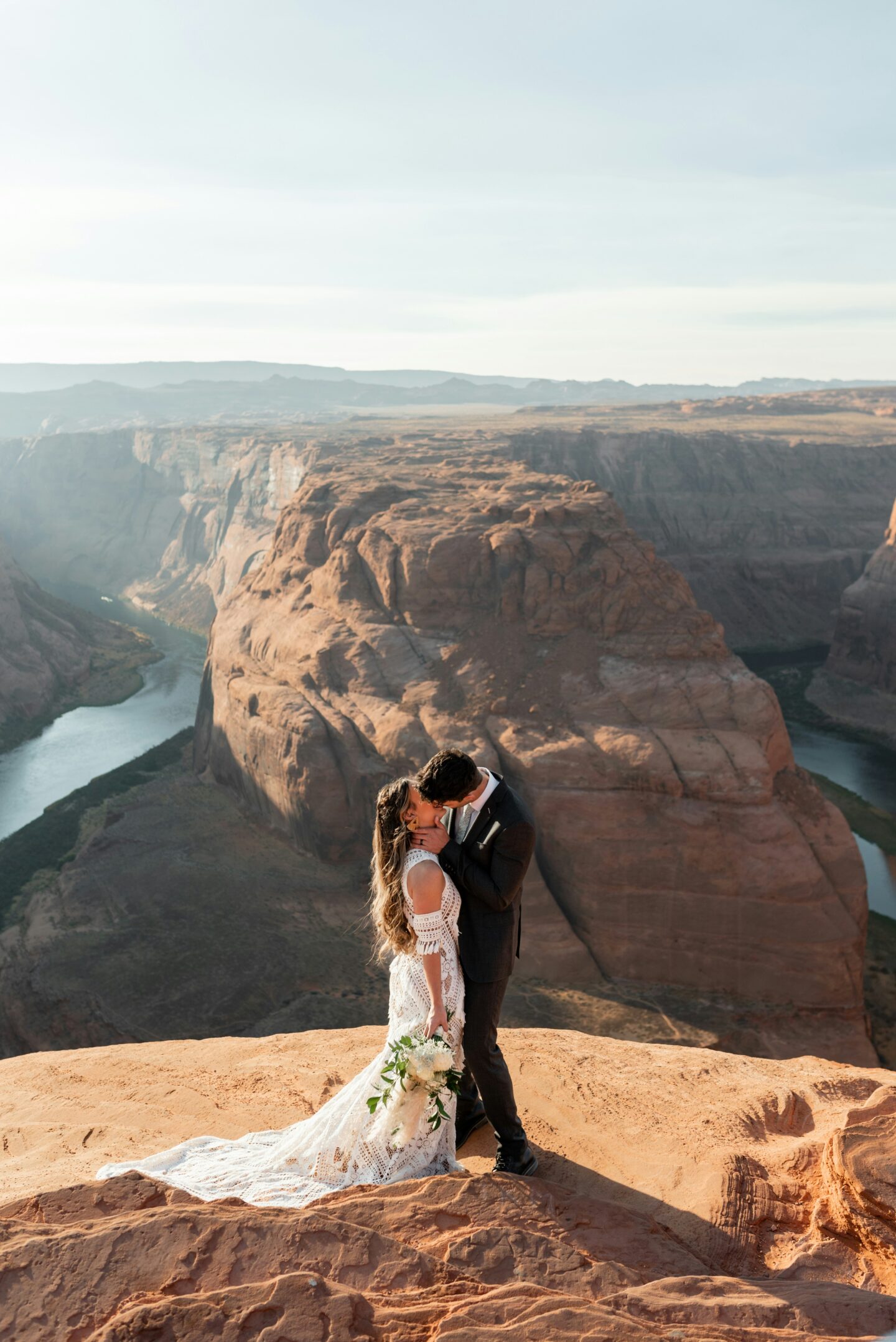 Bride and groom kissing on a cliff overlooking Horseshoe Bend in Arizona, with the Colorado River winding through the canyon behind them.