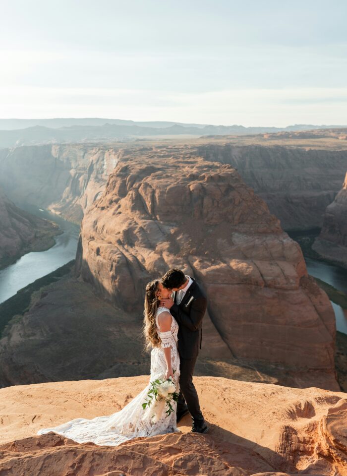 Bride and groom kissing on a cliff overlooking Horseshoe Bend in Arizona, with the Colorado River winding through the canyon behind them.