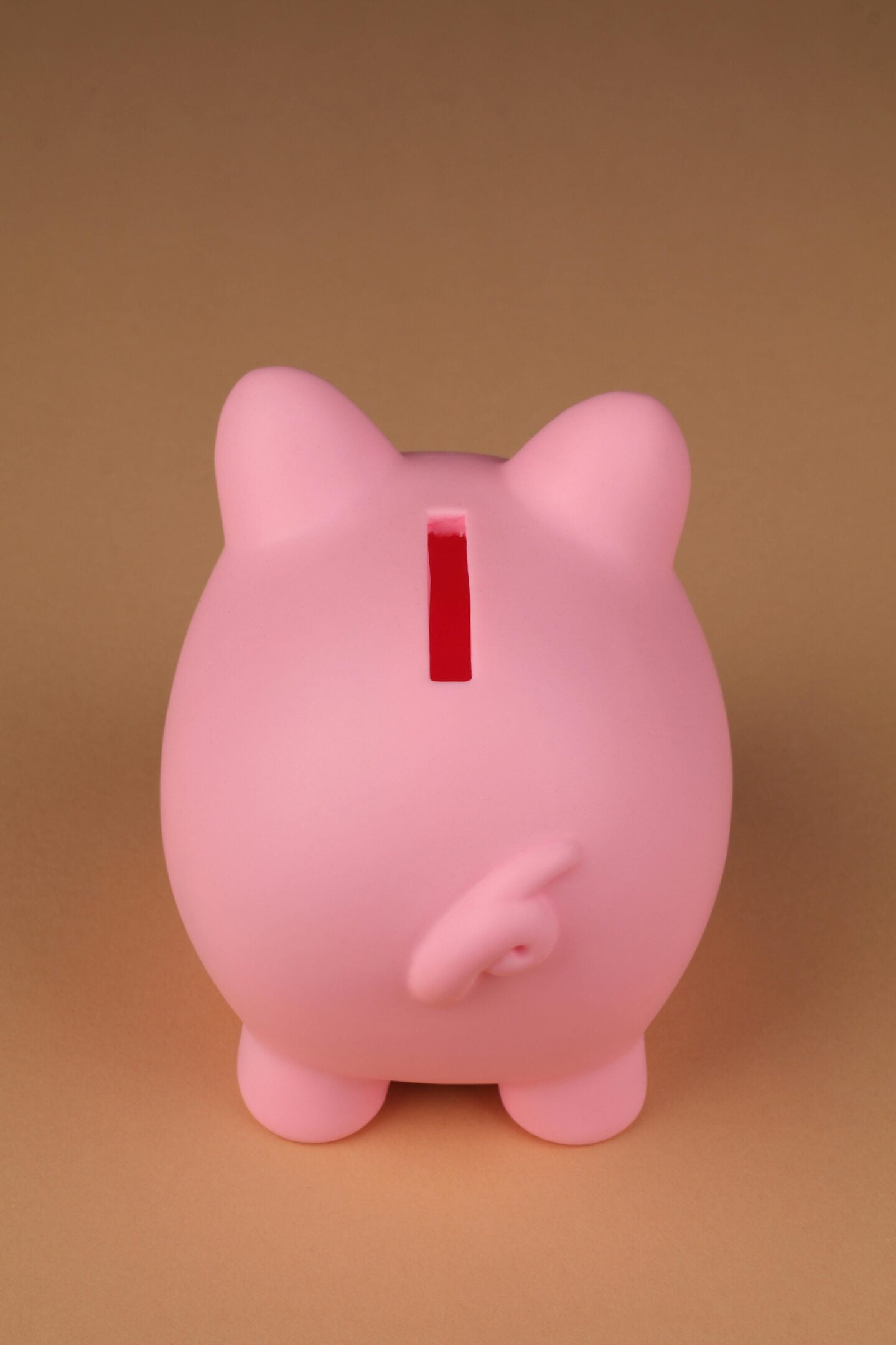 Pink piggy bank viewed from the back with a red coin slot and curly tail, placed against a soft beige background.