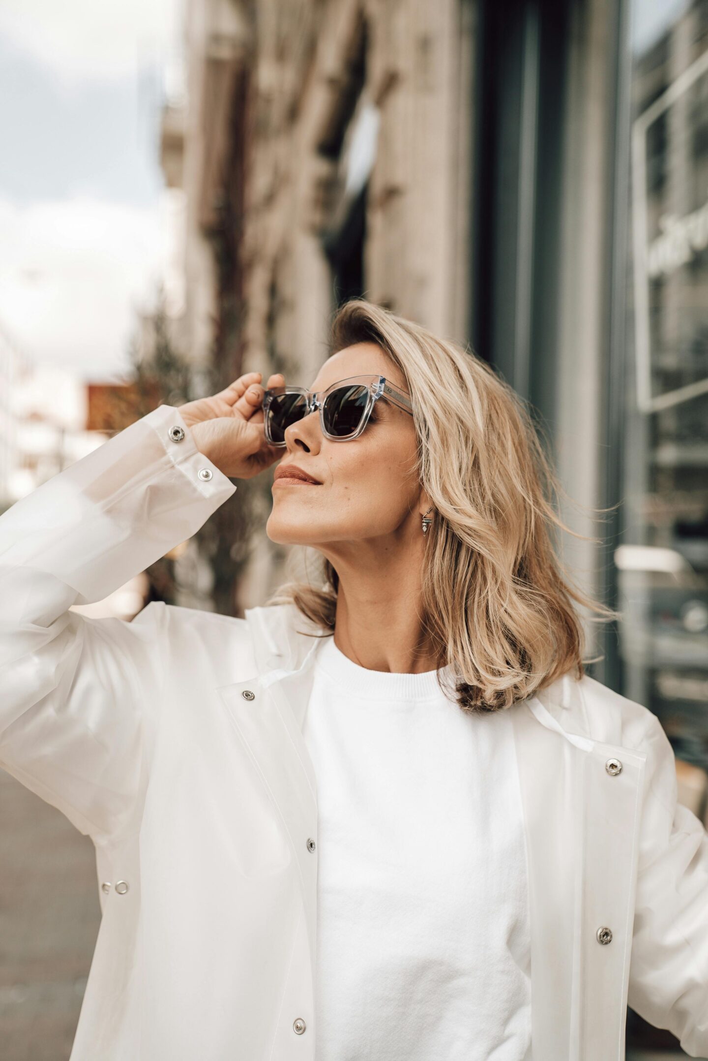 Blonde woman wearing transparent-framed prescription sunglasses and a white jacket standing on a city street, adjusting her glasses in sunlight, modern urban fashion style.