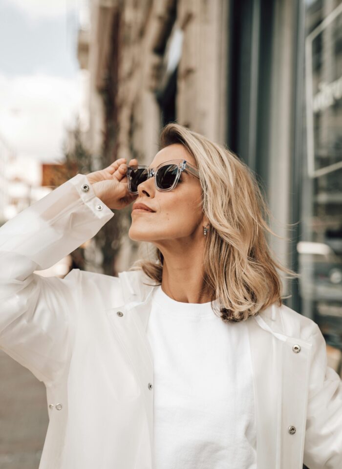Blonde woman wearing transparent-framed prescription sunglasses and a white jacket standing on a city street, adjusting her glasses in sunlight, modern urban fashion style.