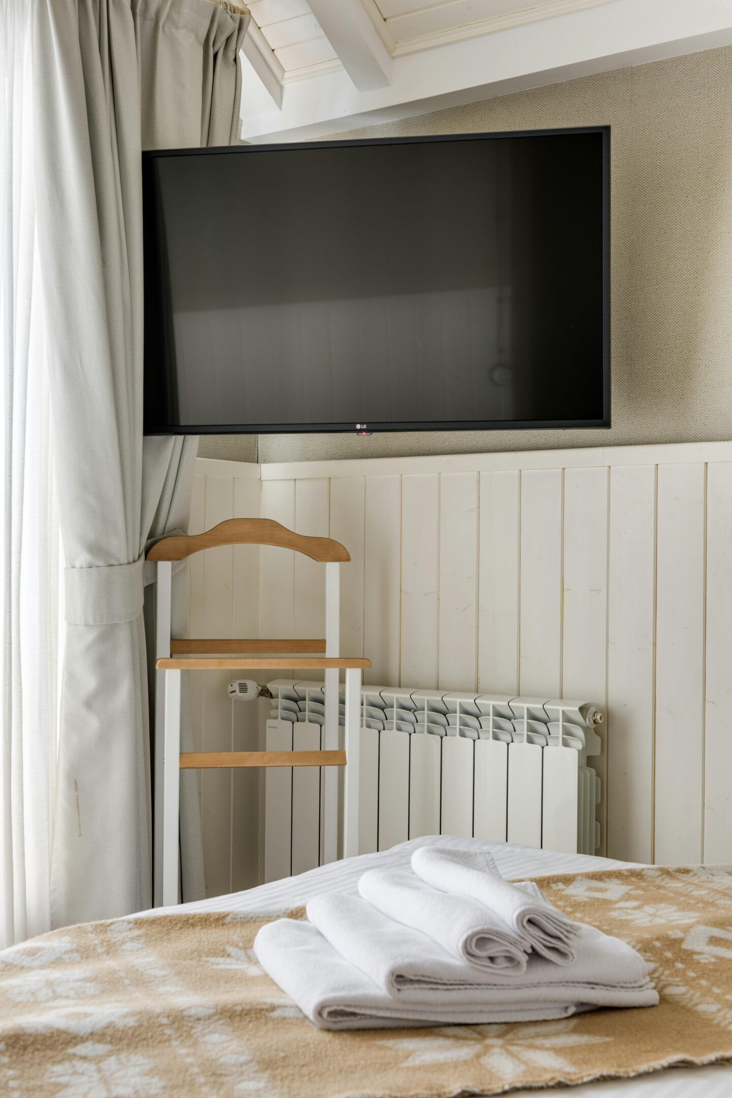 Wall-mounted flat screen TV above a white radiator in a cozy hotel room, with folded white towels on the bed, beige blanket, curtain, and a wooden luggage rack below the television.
