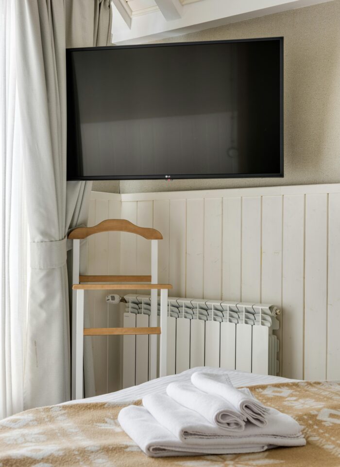 Wall-mounted flat screen TV above a white radiator in a cozy hotel room, with folded white towels on the bed, beige blanket, curtain, and a wooden luggage rack below the television.