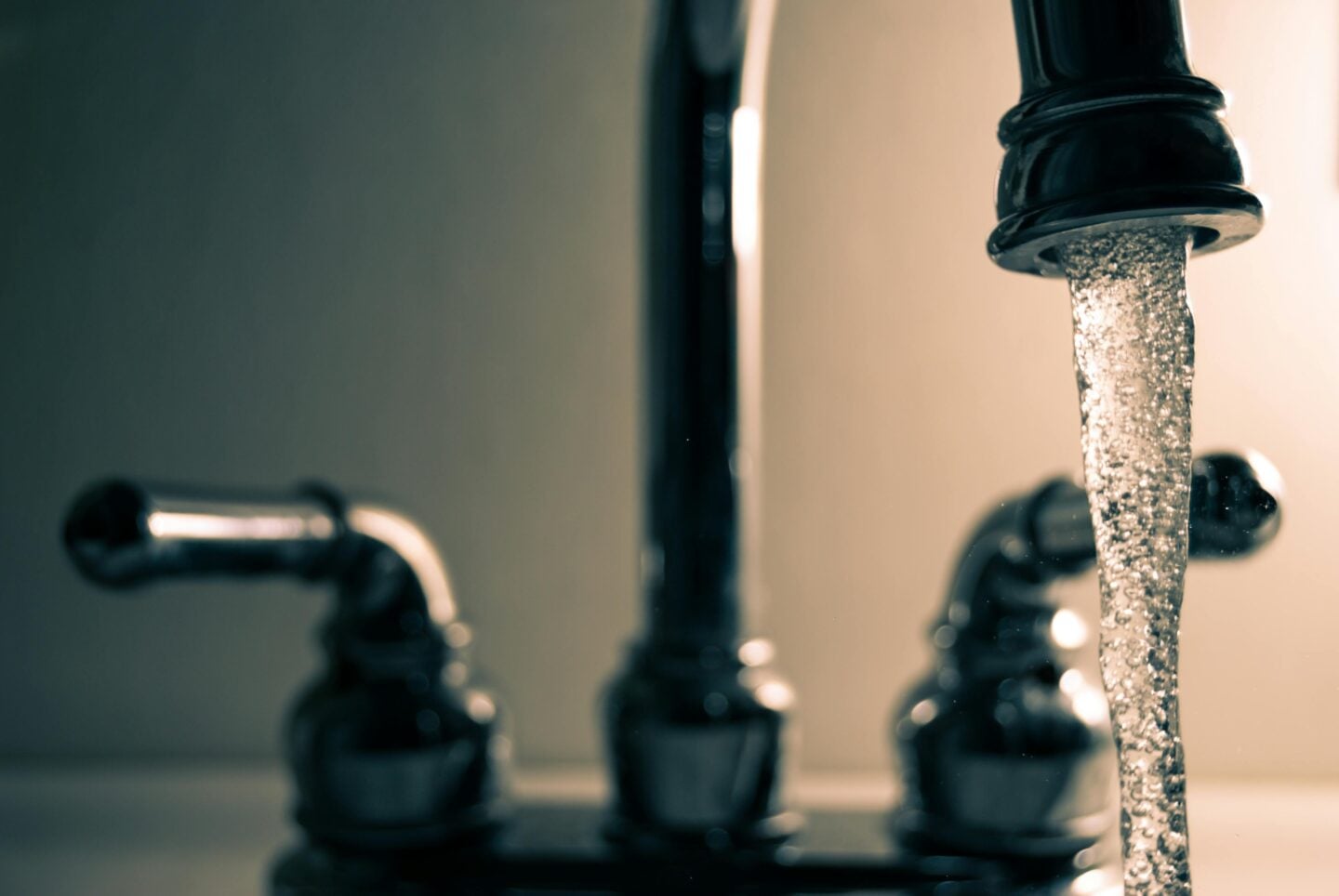Close-up of a bathroom faucet running water, with a steady stream pouring from a dark metal tap while the handles and background remain softly blurred.
